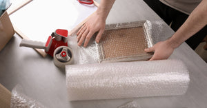 A person wraps a photo frame in bubble wrap on a light gray table with scissors, tape, and packing items nearby.