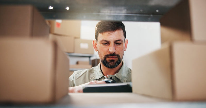 A close-up of a man looking between cardboard boxes on warehouse shelves, with more boxes stacked behind him.