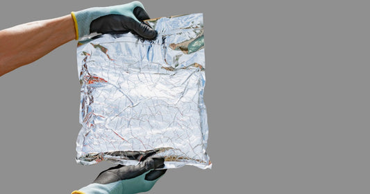 A person wearing gloves holds a foil packaging bag against a gray background, with the material reflecting light.
