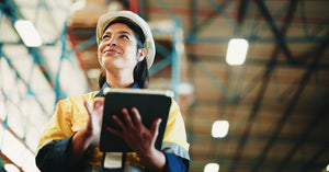 A woman holds a tablet in a warehouse aisle with stacked boxes and tall shelves blurred in the background.