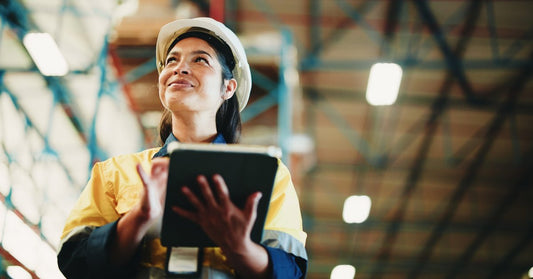A woman holds a tablet in a warehouse aisle with stacked boxes and tall shelves blurred in the background.