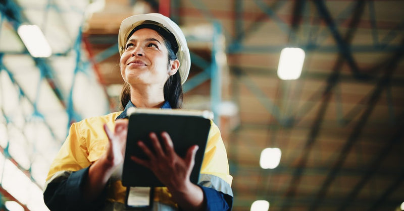A woman holds a tablet in a warehouse aisle with stacked boxes and tall shelves blurred in the background.